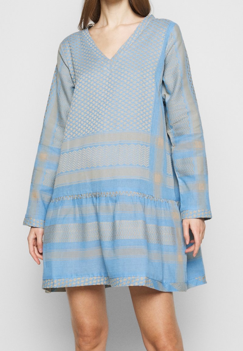 Jasnoniebieska sukienka z dekoltem w kształcie litery V, długimi rękawami i falbanistą spódnicą. Posiada geometryczne i paskowane wzory w kontrastujących odcieniach beżu i niebieskiego.