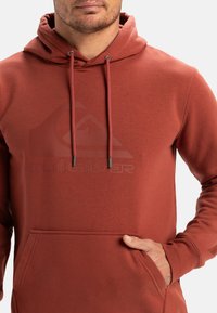 Pánska mikina s kapucňou v hrdzavej červenej farbe z fleecu, s klokankou, nastaviteľnými šnúrkami a tónovým logom na prednej strane. Mäkká textúra.