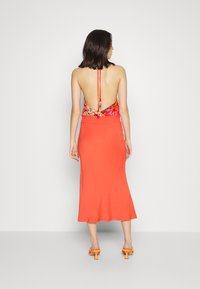 Vista traseira de um vestido de halter com uma parte superior floral vibrante e uma parte inferior sólida em coral, terminado com alças ajustáveis e um silhueta fluida.