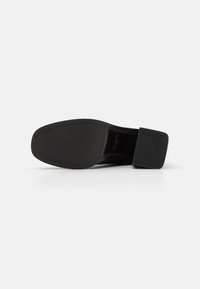 Semelle noire d'une chaussure avec un talon carré reposant sur une surface blanche, montrant des motifs de grip texturés autour des bords.