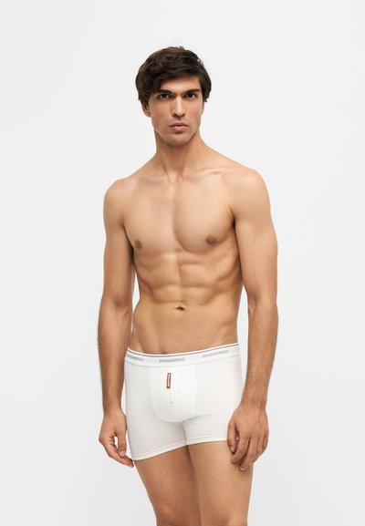 Homme torse nu aux cheveux courts et foncés portant un boxer blanc, debout devant un fond lumineux uni, regardant devant lui avec une expression neutre.