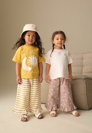 Deux jeunes filles sont debout à l'intérieur, portant un pantalon large à volants, des sandales et des hauts décontractés, l'une avec un bob et de longs cheveux.