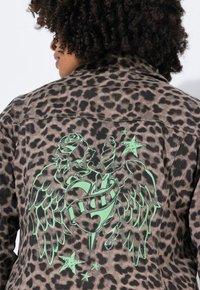 Leopardmönstrad denimjacka med en stor grön grafisk design av en ros, vingar och ett hjärta med en banner, mot en neutral bakgrund.
