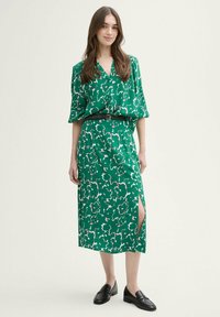 Grünes Blumendruckkleid mit V-Ausschnitt, kurzen Dolmanärmeln, seitlichem Schlitz und tailliertem Gürtel, kombiniert mit schwarzen Loafers.