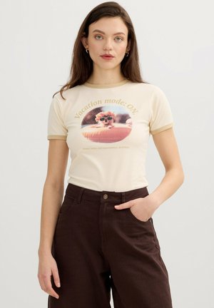 Joven mujer con camiseta crema con texto "Modo vacaciones: ENCENDIDO" y gráfico de perro con gafas de sol, combinado con pantalones de cintura alta color marrón oscuro.
