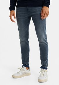 Vaqueros ajustados de mezclilla en azul desgastado, con una textura suave y ligero efecto de desgaste. Combinados con zapatillas blancas de bajo.