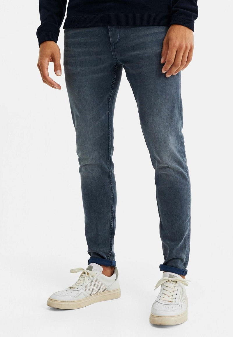 Vaqueros ajustados de mezclilla en azul desgastado, con una textura suave y ligero efecto de desgaste. Combinados con zapatillas blancas de bajo.