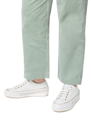 Sneaker bianche con dettagli dorati, parte superiore texturizzata e suola in gomma, abbinate a pantaloni in corduroy verde chiaro, che mostrano un outfit casual.