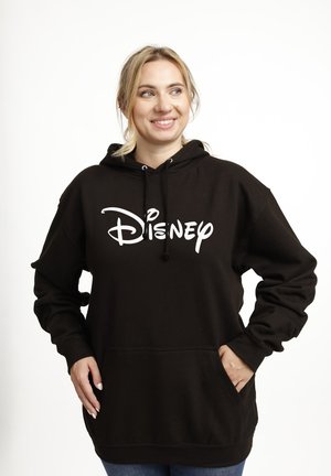 Donna sorridente con capelli biondi indossa una felpa nera con scritta bianca "Disney", in piedi con le mani nelle tasche contro uno sfondo bianco.