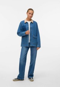 Denim jacket with light tan collar, two front pockets, button closure, and a loose fit. Worn with light blue flared jeans and black sneakers.