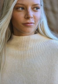 Krémový pletený sveter s vysokým rebrovaným golierom. Mäkka textúra a jemný horizontálny vzor sú viditeľné. Má dlhé, rovné vlasy.