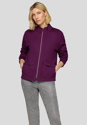 Lila Reißverschlussjacke mit strukturiertem Muster, Stehkragen und zwei Fronttaschen, kombiniert mit grauer Jeans, vor neutralem Hintergrund.