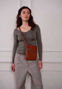 Braune Leder-Umhängetasche mit einer Reißverschluss-Vordertasche und einem abnehmbaren Riemen, die eine glatte Textur und ein minimalistisches Design aufweist.