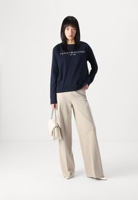 Tengerészkék, hosszú ujjú pulóver fehér "TOMMY HILFIGER EST. 1985" logóval, bézs bővebb nadrággal és krémszínű kézitáskával kombinálva.