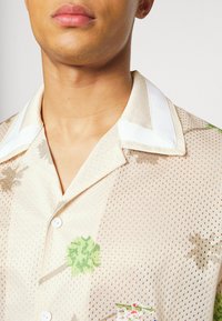 Camisa beige de manga corta con textura de malla. Presenta un cuello blanco, patrones florales en verde y marrón, y un detalle decorativo bordado.