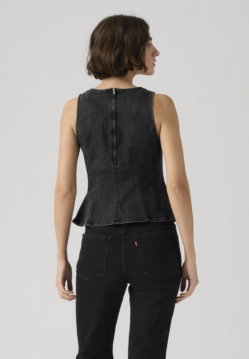 Woman with short brown hair wearing a fitted sleeveless black denim top with a back zipper and black jeans against a plain background.