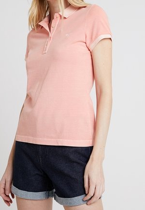 Femme portant un polo rose clair à manches courtes et un short en jean foncé retroussé, debout devant un fond blanc uni.