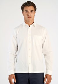 Man staat rechtop met het gezicht naar voren, gekleed in een wit overhemd met lange mouwen en donkere broek, tegen een effen achtergrond.