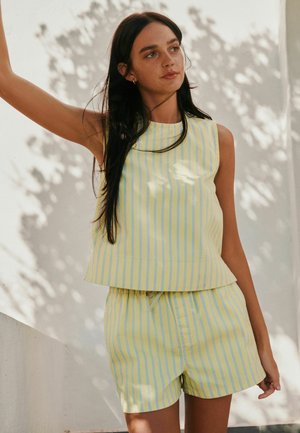 Femeie cu păr lung și închis la culoare, purtând un top galben fără mâneci și pantaloni scurți cu dungi albastre verticale, stând lângă un perete alb iluminat de soare.