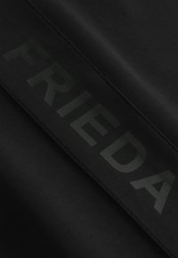 Czarna tkanina o gładkiej fakturze z dużym, przytłumionym zielonym napisem "FRIEDA" wzdłuż poziomego pasa. Widoczne detale szycia.