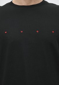 T-shirt noir à col rond avec des lettres rouges espacées formant H U G O sur la poitrine.