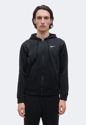 Homme aux cheveux foncés mi-longs portant un sweat à capuche zippé noir Nike et un pantalon noir, debout devant un fond clair uni.