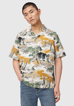 Mand med kort hår iført en kortærmet skjorte med knapper med grøn, gul og sort natur- og dyreprint, kombineret med blå jeans.