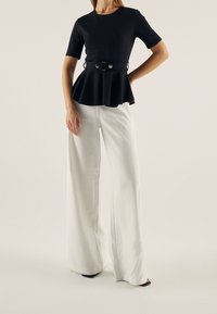 Černý peplum top s krátkými rukávy, stažený v pase s kovovou sponou. Kombinováno s bílými širokými kalhotami a černými podpatky. Jednoduché pozad�í.