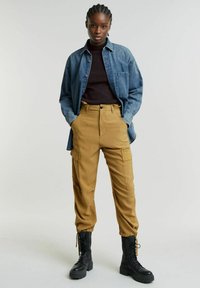 Camicia di jeans, maniche lunghe, azzurro chiaro; maglia nera a maniche lunghe; pantaloni cargo cammello con tasche; stivali combat neri con lacci.