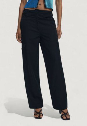 Woman wearing wide-leg black cargo pants and black strappy heeled sandals standing against a plain background.