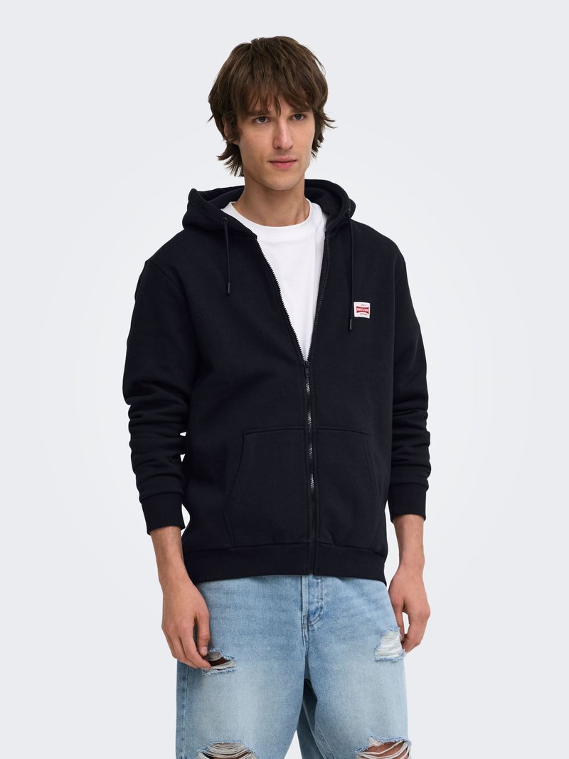 Jeune homme portant un sweat à capuche noir zippé avec un petit logo, un t-shirt blanc et un jean bleu clair déchiré, debout devant un fond uni.
