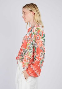 Blumenbluse in Orange mit grünen, rosa und schwarzen Mustern. Lockere Passform, Puffärmel, glatte Textur. Keine sichtbaren Verschlüsse.