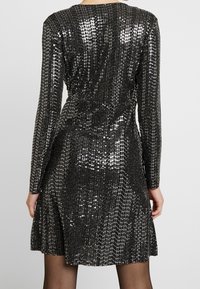 Svart paljettklänning med långa ärmar, figursydd liv och utsvängd kjol. Skimrande textur med en slät yta och anmärkningsvärda reflexdetaljer.