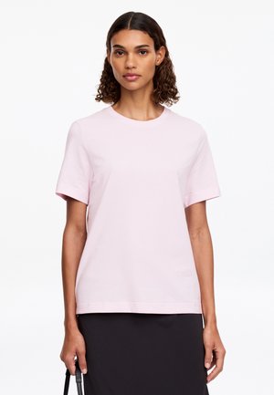 Femme aux cheveux bouclés mi-longs, portant un t-shirt rose clair à manches courtes et une jupe noire, tenant une anse de sac noire.