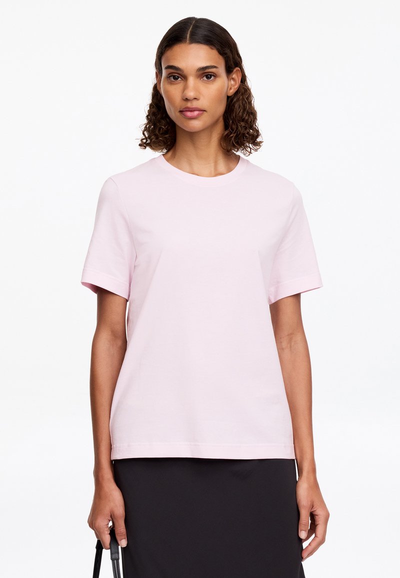 Femme aux cheveux bouclés mi-longs, portant un t-shirt rose clair à manches courtes et une jupe noire, tenant une anse de sac noire.