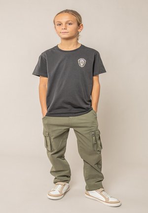 Jeune personne debout les mains dans les poches, portant un t-shirt graphique foncé, un pantalon cargo olive et des baskets blanches sur un fond neutre.