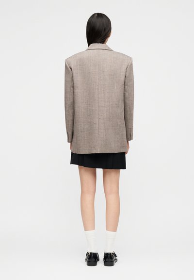 Femme aux longs cheveux bruns vue de dos portant un blazer beige surdimensionné, une jupe noire, des chaussettes blanches et des mocassins noirs sur un fond blanc.