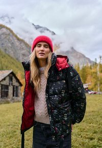 Czarna kurtka puchowa z kwiatowym wzorem, czerwoną podszewką i kieszeniami na zamek; noszona na jasnobeżowym swetrze z dzianiny i czarnych dżinsach. Czerwona dzianinowa czapka.