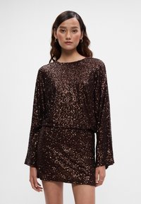 Haljina od bronze s cirkonima, dugih rukava, okruglog izreza i prilagođenog kroja. Materijal ima sjajan, teksturiran finiš.