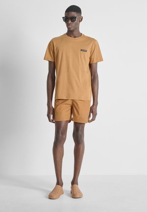 Homme portant un t-shirt et un short beige assortis, des chaussures beige sans lacets et des lunettes de soleil noires, debout devant un fond clair uni.