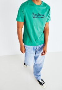 Camiseta de algodón verde con cuello redondo y mangas cortas, presenta un logo impreso en azul marino "Pepe Jeans" en la parte frontal. Se lleva con pantalones vaqueros de tono azul claro.