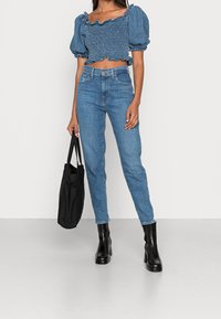 Top en denim avec des manches bouffantes et des bords festonnés, associé à un jean bleu taille haute à jambe droite et des bottines noires à talons. Sac fourre-tout noir.