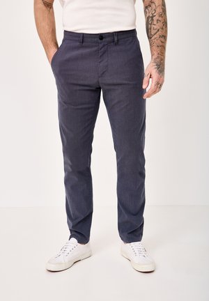 Man wearing dark gray slim-fit pants, white sneakers, and a white shirt with tattooed forearms visible.