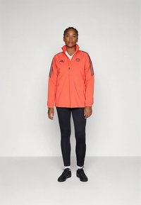 Lightweight orange athletic jacket with black stripes, zip closure, and logo on chest, paired with black leggings and black sneakers.
