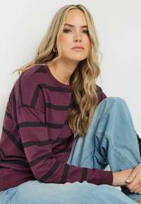 Felpa a maniche lunghe a righe marrone e nera con polsini a coste, abbinata a jeans oversized blu chiaro. Modella seduta, che guarda verso la macchina fotografica.