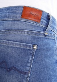 Pantalones vaqueros de denim azul con una etiqueta de cuero marrón que dice "Pepe Jeans London", remaches metálicos y un patrón de costura ondulada en el bolsillo trasero.