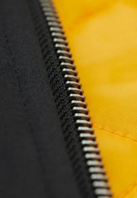 Schwarzer Reißverschluss auf einem leuchtend gelben Stoff, der metallische Zähne und strukturiertes schwarzes Material neben der glatten gelben Oberfläche zeigt.