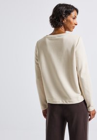 Femme aux cheveux courts et bouclés portant un pull beige à manches longues et un pantalon sombre, de profil contre un fond gris clair uni.
