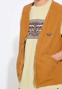 Personne portant un gilet beige avec une fermeture éclair sur un t-shirt graphique beige avec des motifs abstraits de visages roses et noirs. Main dans la poche du gilet.