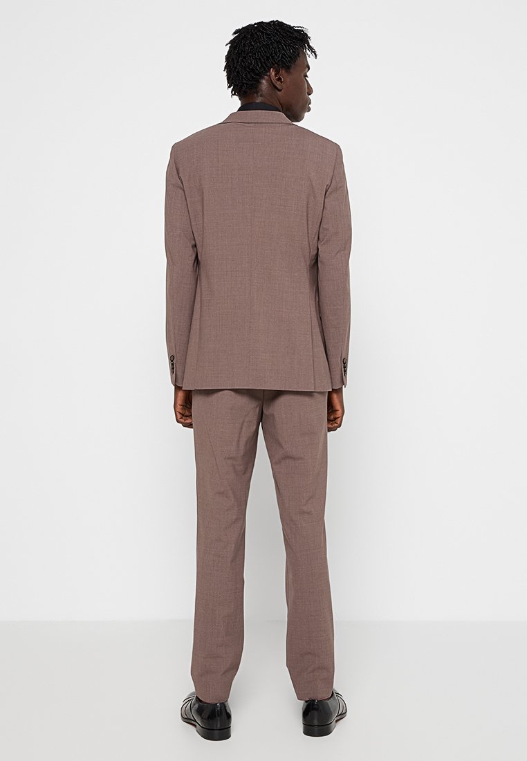 Uomo in piedi, girato di spalle, che indossa un completo mauve con pantaloni coordinati e scarpe eleganti nere, su uno sfondo bianco.